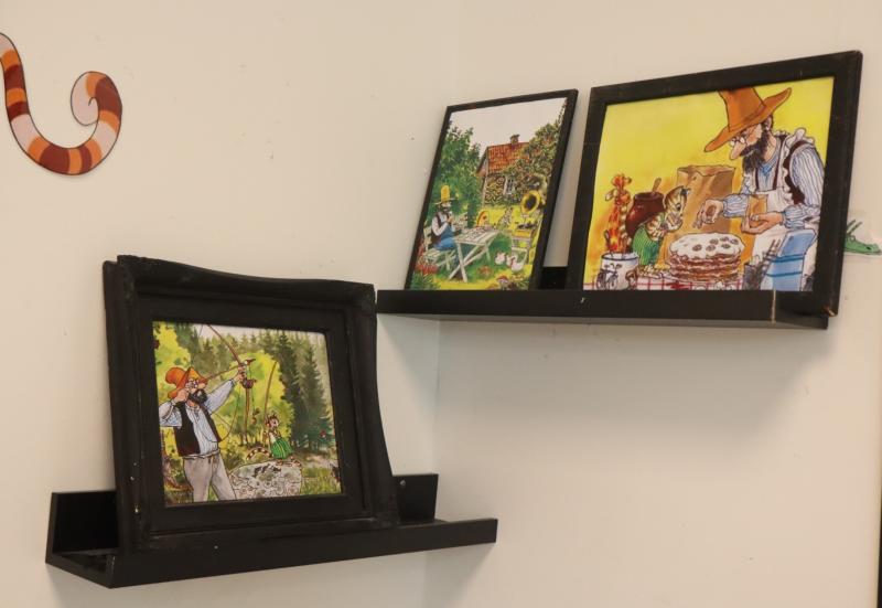 På två svarta hyllor på en vit vägg finns färgglada illustrationer i svarta ramar; en ram på den nedre hyllan är synligt böjd och skadad.