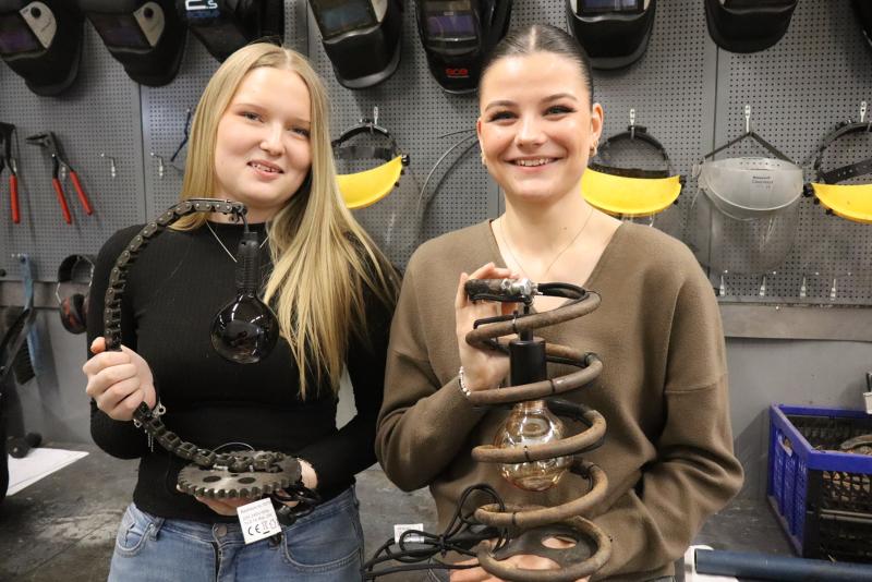 Två unga kvinnor står i en verkstad och håller var sin lampa gjord av återanvända mekaniska delar, med svetshjälmar och verktyg synliga i bakgrunden.