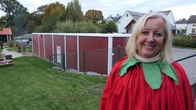 En kvinna i röd dräkt med grön krage står utomhus på gräset framför en röd byggnad med staket, med hus och träd i bakgrunden.