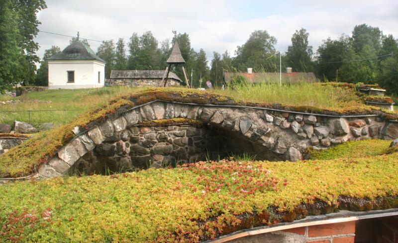 En stenbåge täckt av gräs och mossa, med en liten vit byggnad, träd och andra strukturer i bakgrunden under en molnig himmel.