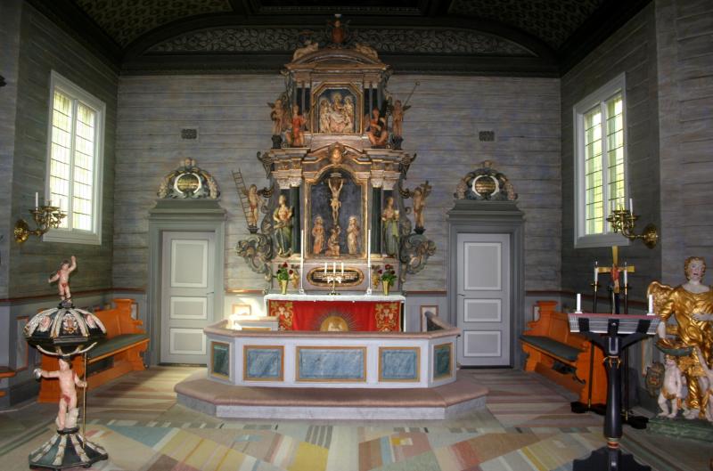 Ett kyrkoaltare med utsmyckade religiösa skulpturer, ljus och en röd duk, omgivet av träbänkar och dekorerade väggar med fönster på vardera sidan.