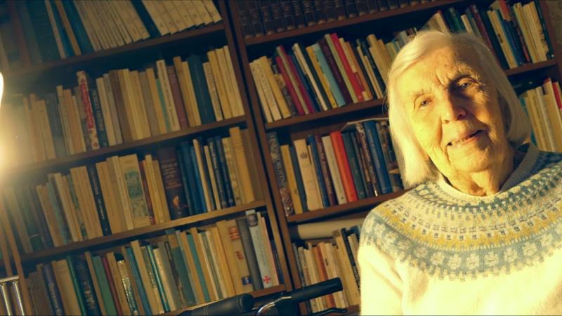 Äldre kvinna med vitt hår och mönstrad tröja sitter framför bokhyllor fyllda med böcker, upplysta av ett starkt ljus till vänster i bild.