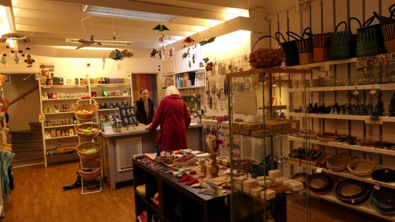 En kund står vid disken och pratar med en anställd i en väl upplyst butik som säljer hantverk, korgar, smycken och olika handgjorda föremål som visas på hyllor och bord.