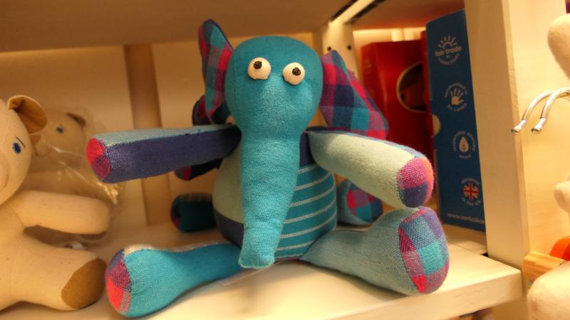 En blå och turkos gosedjurselefant med rutmönstrade öron och lemmar står på en hylla bland andra gosedjur.