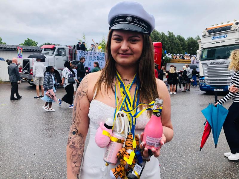 En ung kvinna som bär en examensmössa och vit klänning står utomhus. Hon har olika föremål hängande från hennes hals, håller i en rosa drink och har synliga tatueringar. Lastbilar och människor står i bakgrunden.