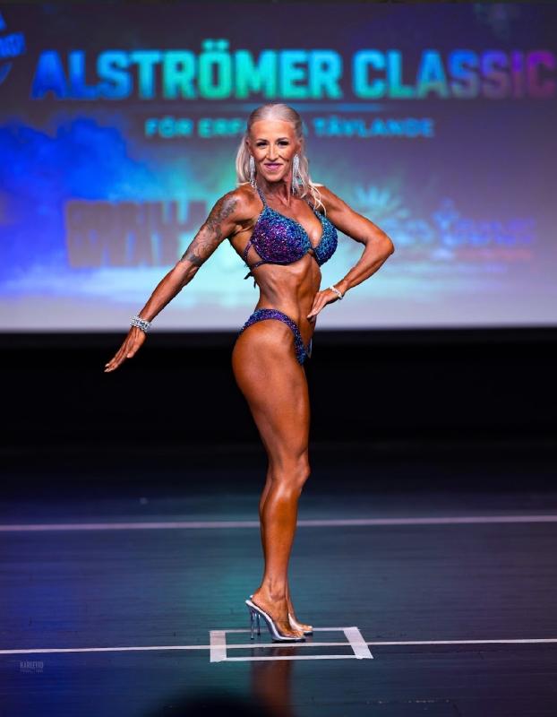En kvinnlig kroppsbyggare i en glittrande bikini och högklackade skor poserar på scenen vid en kroppsbyggartävling som heter Alströmer Classic.