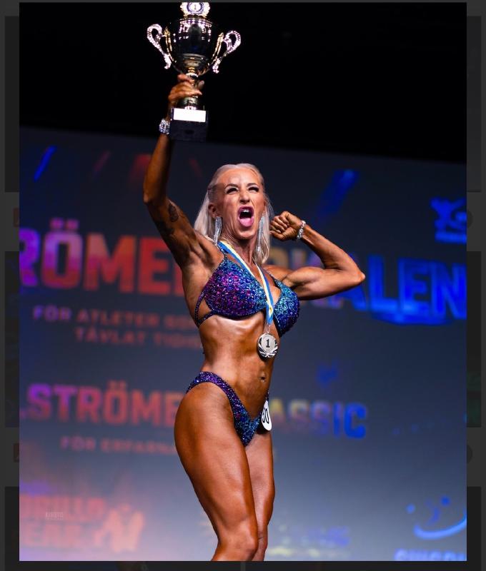 En kvinnlig kroppsbyggare i en glittrande bikini poserar på scenen med en pokal högt i luften, iklädd en medalj och tävlingsnummer 20, och firar sin seger.