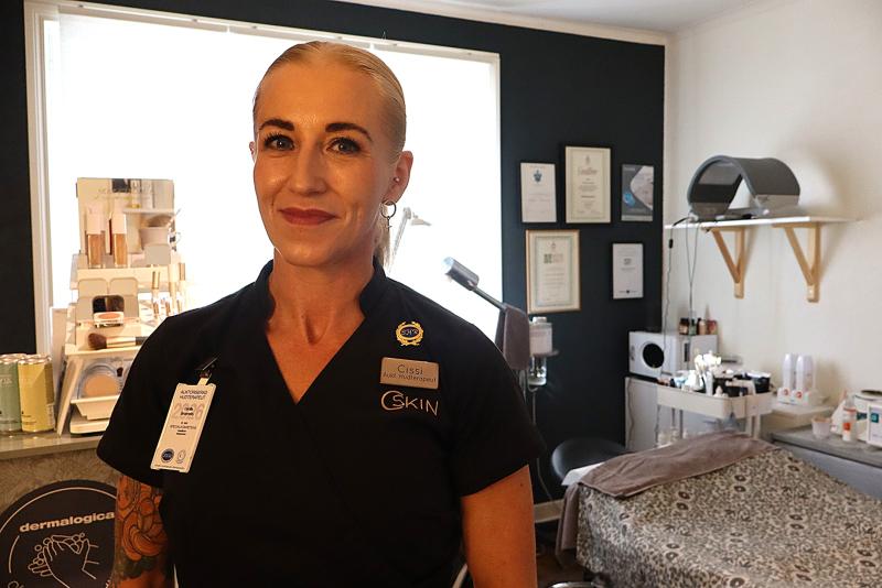 En kvinna i svart uniform med namnskylt står i ett väl upplyst behandlingsrum för hudvård, där utrustning, certifikat och produkter syns tydligt.