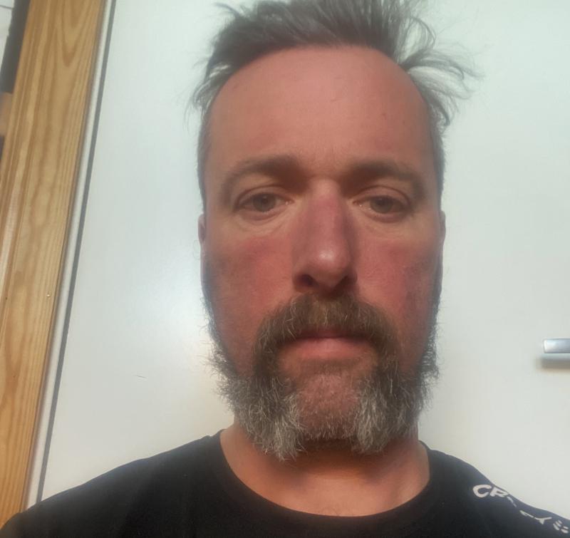 En man med kort hår och skägg står vänd mot kameran, iklädd en svart skjorta och står framför en trädörr och vit vägg.