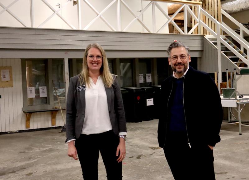 Två personer står inomhus på ett grått betonggolv. Kvinnan till vänster bär glasögon och en grå kavaj. Mannen till höger bär glasögon och mörk jacka. De är i en industriell miljö.