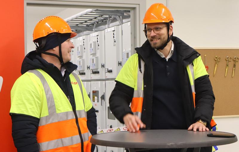 Två män i orange skyddshjälm och reflexväst står och pratar vid ett runt bord i en industrimiljö med kontrollpaneler och verktyg i bakgrunden.