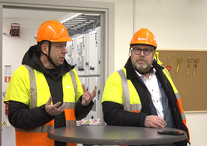 Två män i varseljackor och orangea skyddshjälmar står inomhus, den ene gestikulerar medan han talar och den andre lyssnar vid ett runt bord.