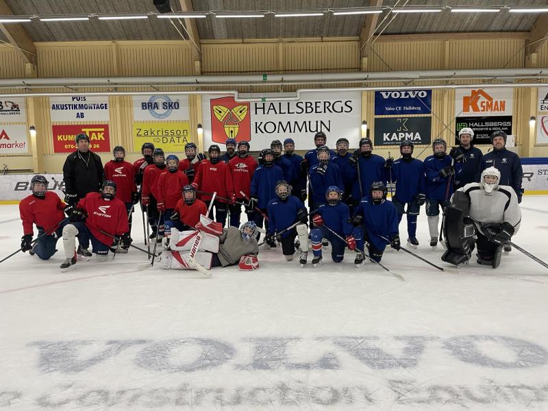 En grupp hockeyspelare i röda och blåa tröjor poserar på en ishall inomhus. Några spelare står, andra knäböjer eller sitter på isen. Hockeyutrustning och klubbor är synliga.