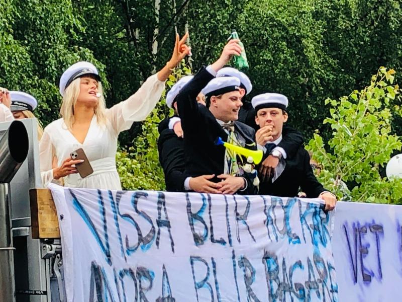 En grupp unga vuxna i högtidlig klädsel och vita kepsar firar på ett flöte och håller i drinkar och en banderoll med svensk text. Träden syns i bakgrunden.