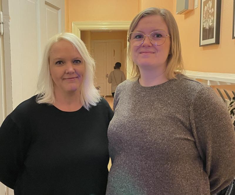 Två personer med ljust hår står inomhus framför en hall. En person bär en svart topp, och den andra bär glasögon och en grå topp. Konst visas på de persikofärgade väggarna.