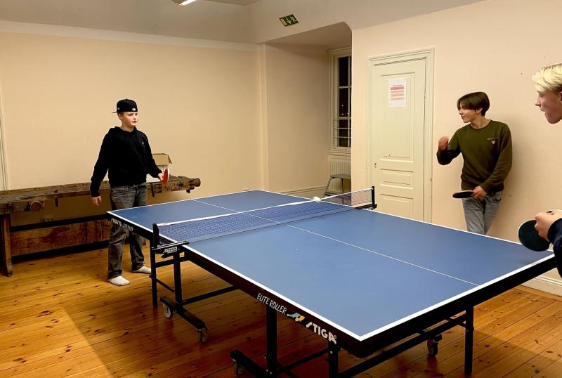 Tre personer som spelar bordtennis i ett rum med trägolv och beiga väggar. En person serverar medan två andra förbereder sig för att spela.