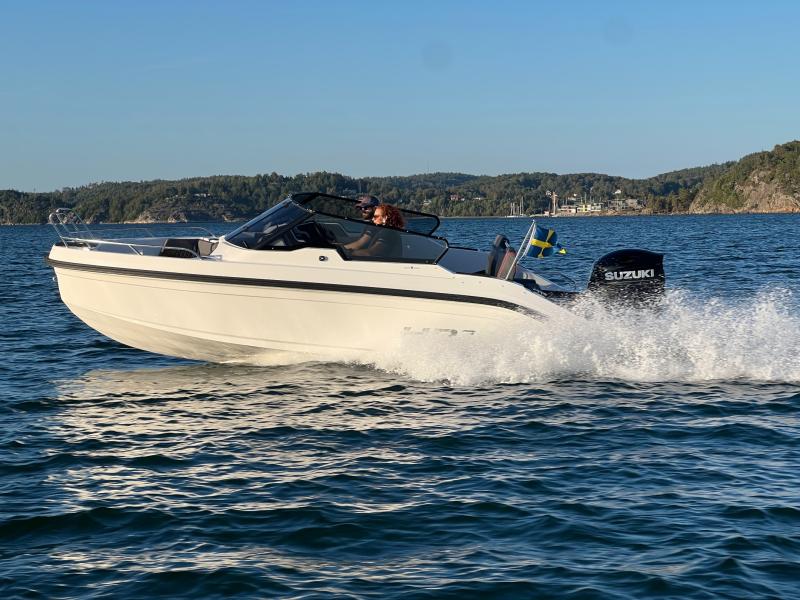 En vit motorbåt med en Suzuki utombordsmotor kör över ett lugnt vattendrag med en skogsklädd strandlinje i bakgrunden.