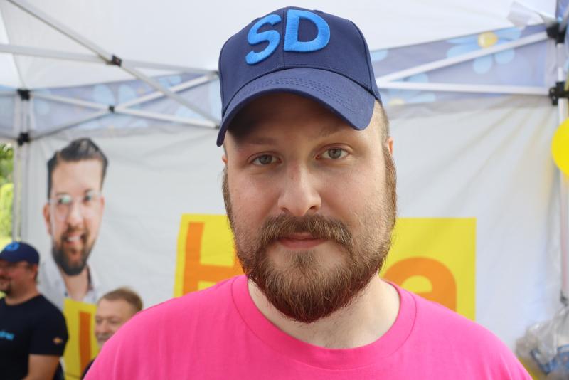 En skäggig man med en blå keps med bokstäverna ”SD” och en rosa skjorta står framför en monter med en gul skylt och en affisch av en annan man i bakgrunden.