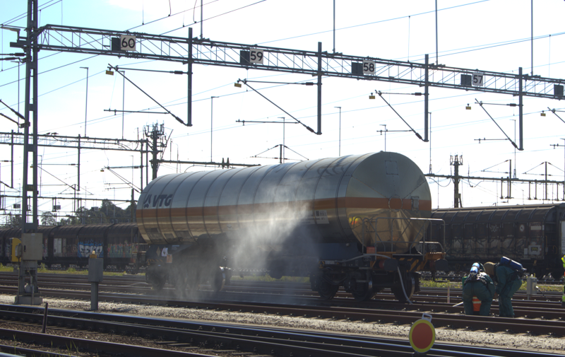 En stor cylindrisk tankvagn avger ånga på tågspår, med arbetare i skyddsutrustning som står i närheten. Kontaktledningar och numrerade skyltar är synliga.