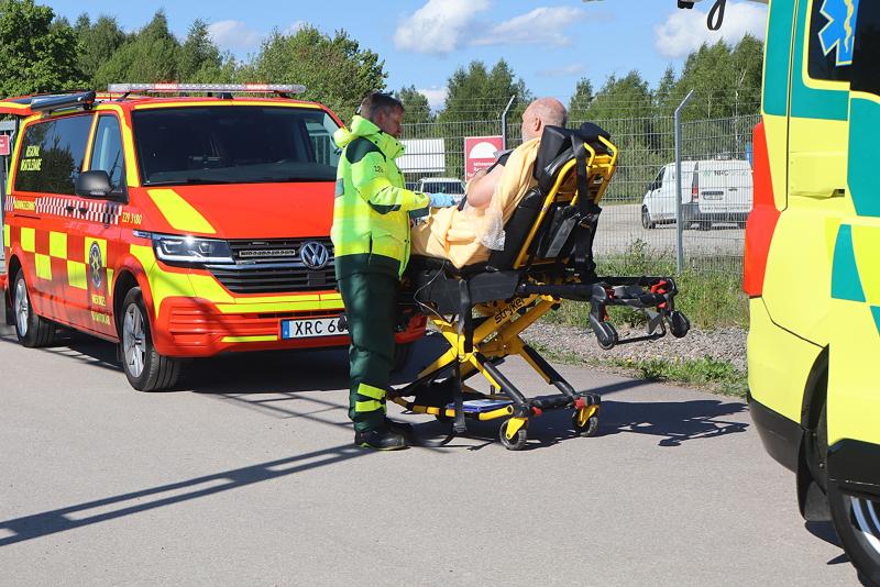 En ambulanssjukvårdare hjälper en sittande person på en bår bredvid en ambulans och ett rött utryckningsfordon en solig dag.