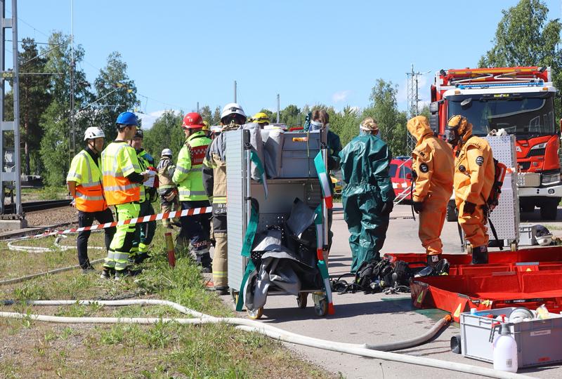 Räddningspersonal, vissa i skyddsdräkter, samlas i närheten av utrustning och en brandbil vid en utomhusplats en solig dag.