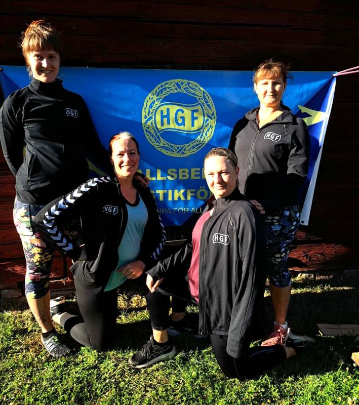 Fyra kvinnor i träningskläder poserar utomhus på gräset framför en blå HGF-banderoll, med leenden och avslappnade kroppshållningar.