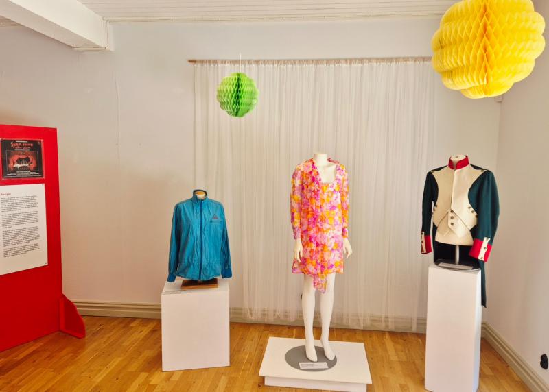 Tre skyltdockor visar upp en blå jacka, en rosa blommig klänning och en jacka i historisk stil i en minimalistisk museutställning med informationstexter och hängande pappersdekorationer.