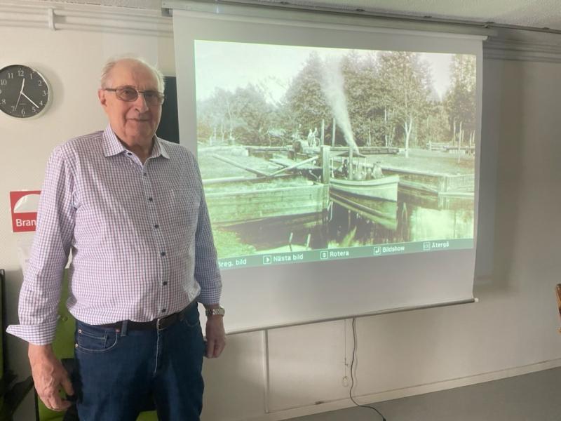 En äldre man står bredvid en projektorduk och visar en historisk svart-vit bild av ett lås och en pråm. En väggklocka och en brandskyddsskylt syns i bakgrunden.