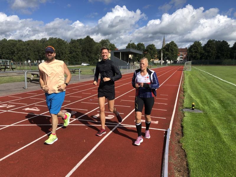 Tre personer joggar på en röd löparbana bredvid en gräsbevuxen plan, under en delvis molnig himmel. En man i blå shorts, en man i svart och en kvinna i atletisk kläder tränar tillsammans.