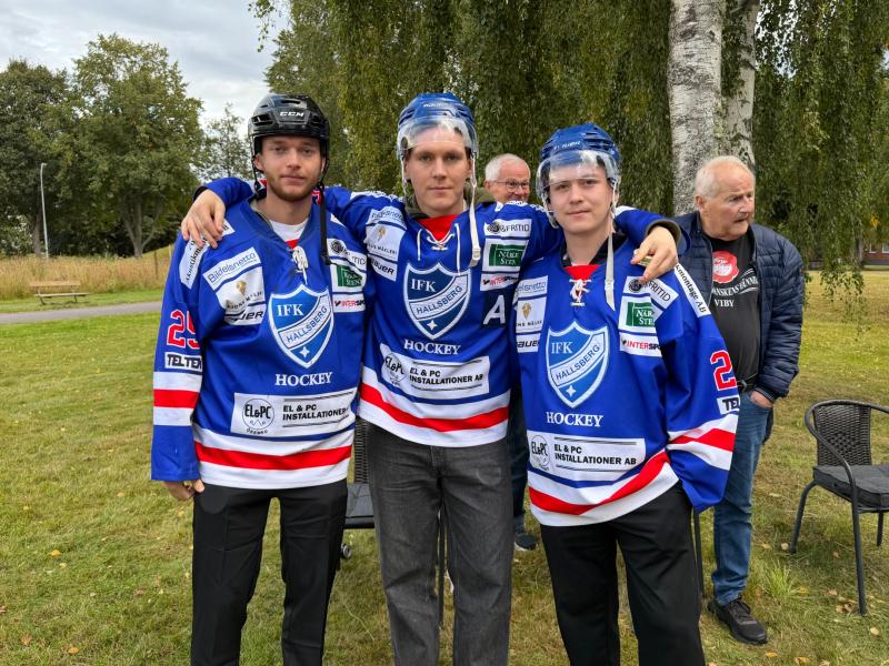 Tre unga ishockeyspelare i blå och röda IFK-tröjor står arm i arm på gräset, med flera äldre män och träd i bakgrunden.