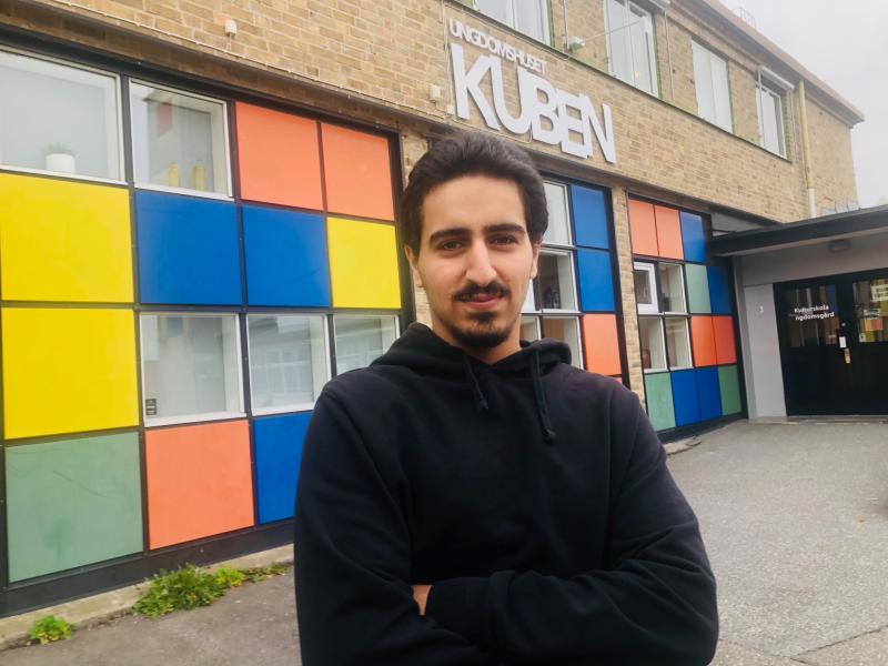 En man med armarna i kors står utanför en byggnad med färgglada fyrkantiga paneler och en skylt med texten 