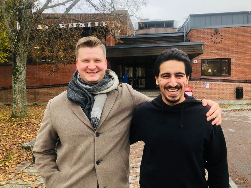 Två män står utanför en tegelbyggnad en höstdag och ler mot kameran; den ene har armen runt den andres axel.