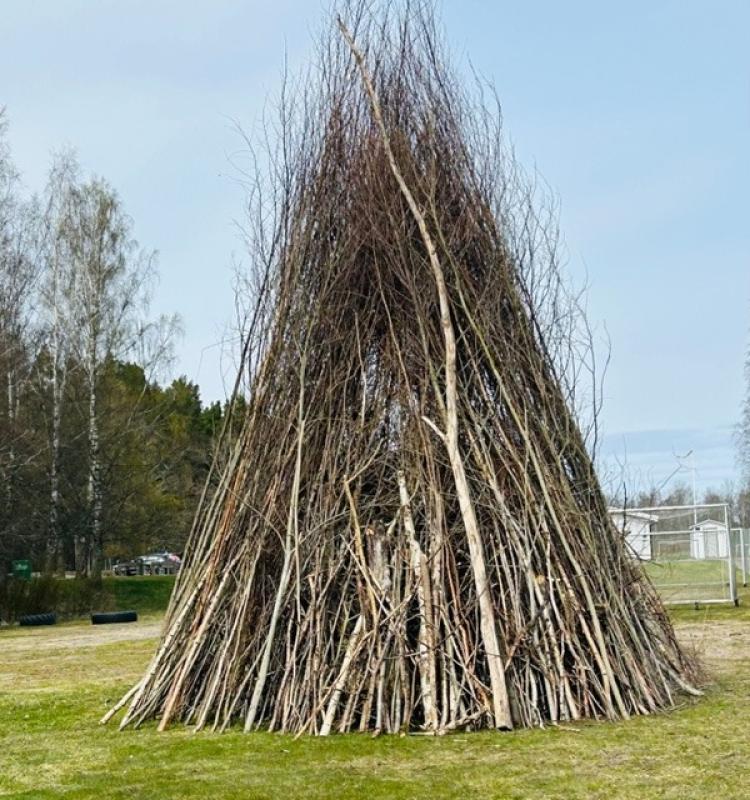 En stor utomhuskonstruktion bestående av upprättstående trägrenar som är placerade i en konisk, tipi-liknande form på en gräsmatta.