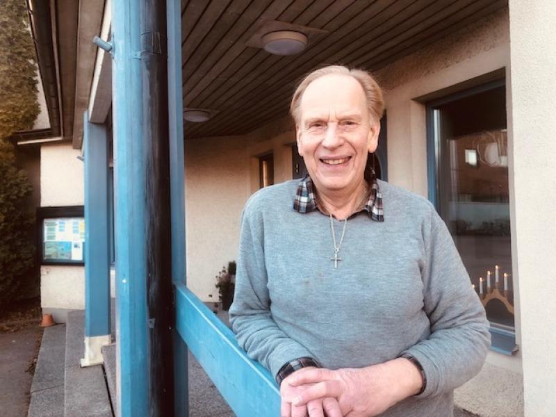 En äldre man i grå tröja står utanför en byggnad med blå balkar och ler mot kameran.