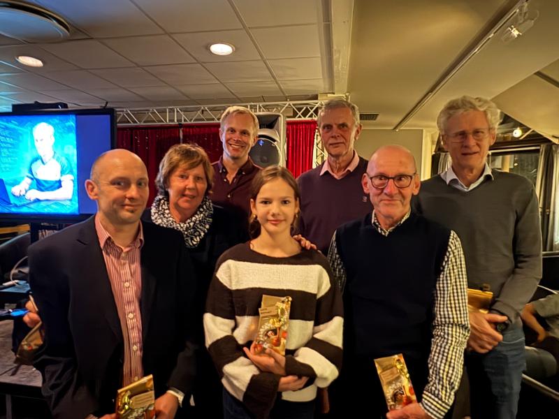 En grupp på sju personer, inklusive ett barn, står inomhus och håller i vad som verkar vara priser eller gåvor och poserar för ett foto. En skärm och röda gardiner syns i bakgrunden.