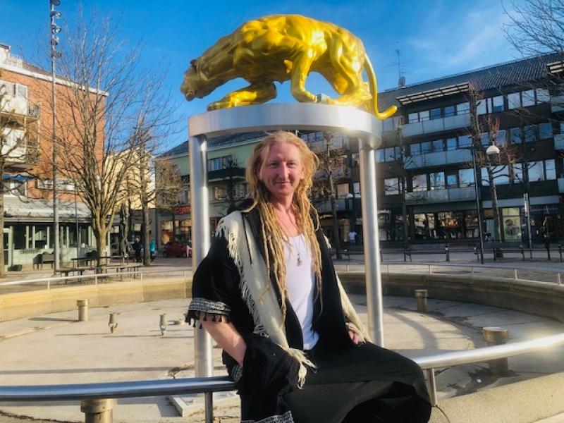 En person med långa blonda dreadlocks sitter på ett räcke framför en staty av den gyllene pantern på ett torg i staden en solig dag.
