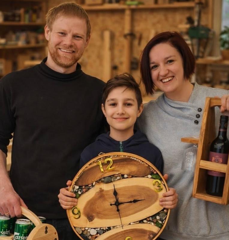 Tre personer står i en verkstad och ler. Barnet håller i en träklocka, mannen håller i en trätråg med burkar och kvinnan håller i en vinhållare i trä med en flaska.