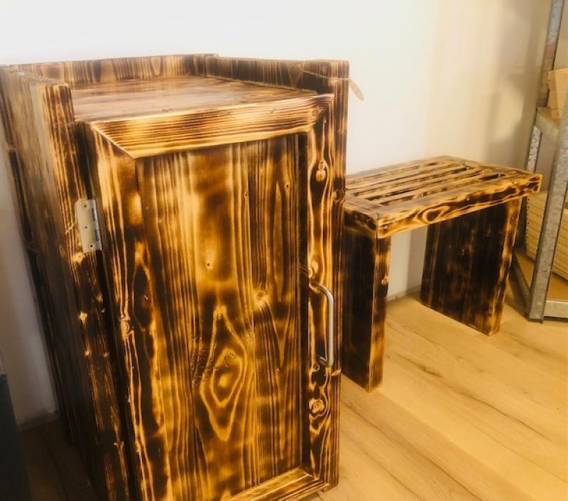 Ett träskåp med gångjärnsdörr och en matchande bänk, båda med bränd träfinish, står på ett ljust trägolv mot en vit vägg.