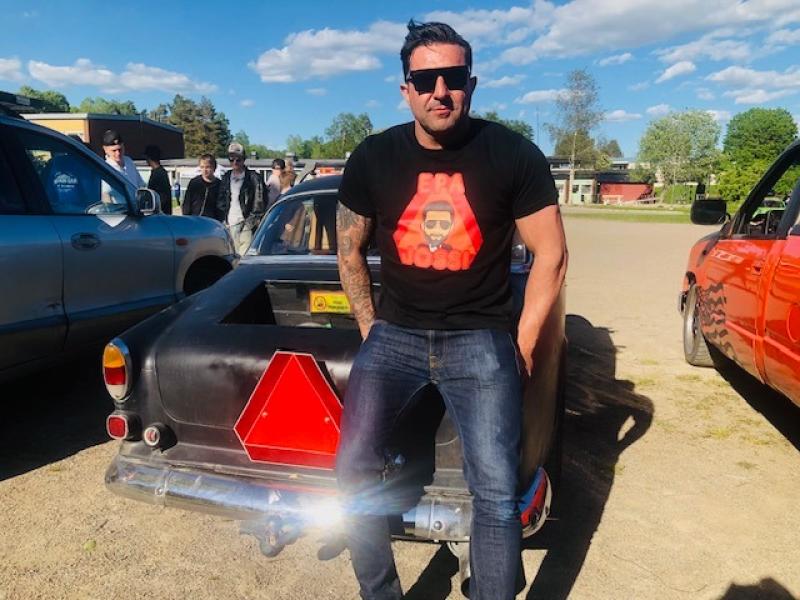 En man i solglasögon och svart t-shirt står framför en klassisk bil med en röd reflekterande triangel fäst på bakluckan, utomhus en solig dag.