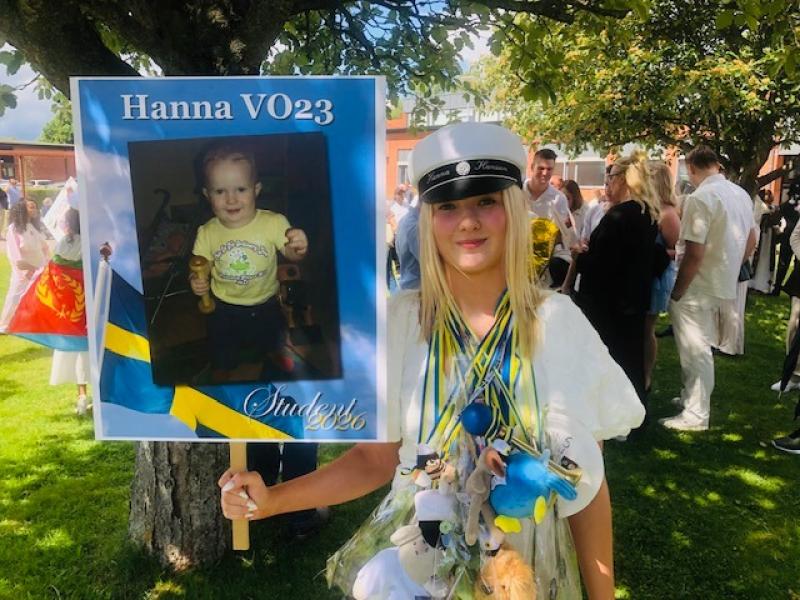 En ung kvinna i vit studentmössa står utomhus och håller en skylt med ett babyfoto, svenska flaggor och 