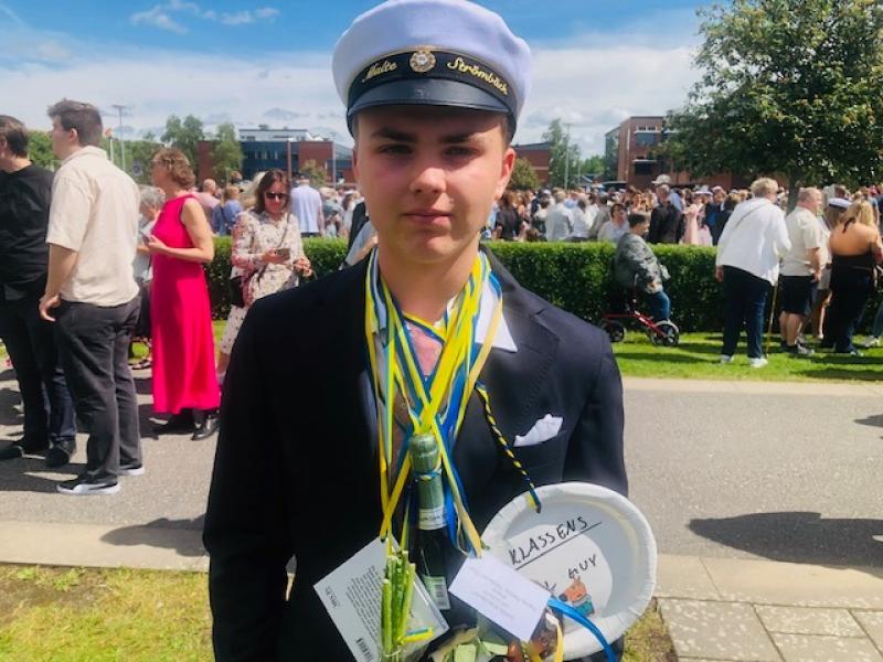 Ung person i studentmössa och mörk kavaj står ute bland en folkmassa, med presenter och en dekorerad tallrik runt halsen, troligen vid en examensfest.