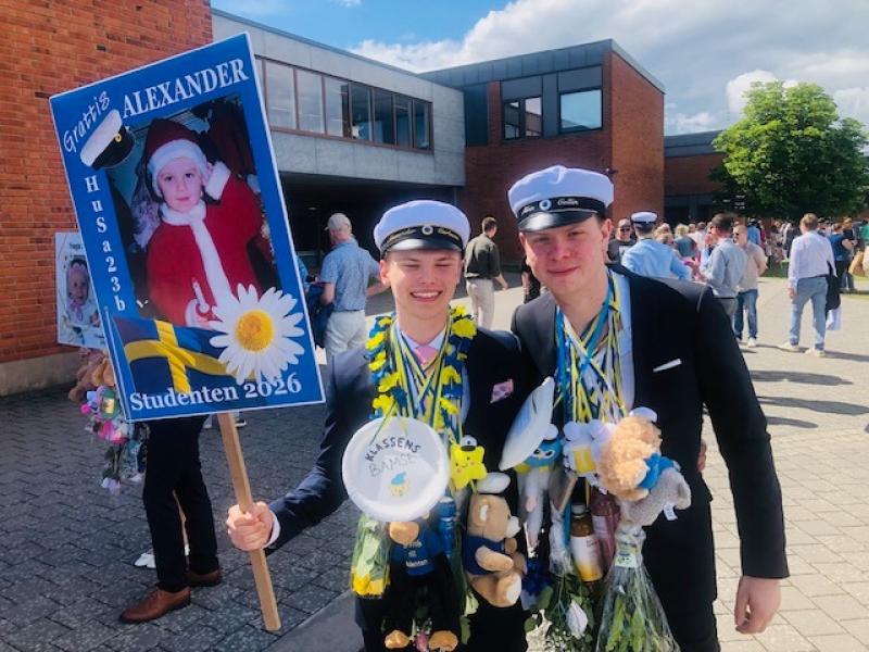 Två unga män i kostym och studentmössa står utanför en byggnad; den ene håller i en dekorerad examensskylt med ett barndomsfoto och 