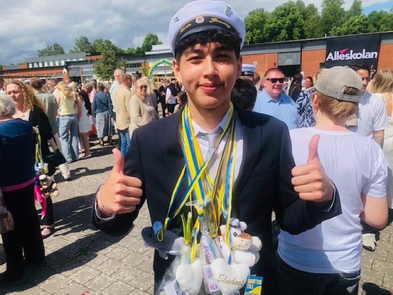En ung man i studentmössa och kostym ger två tummar upp, med band och leksaker runt halsen, bland en folkmassa utomhus.