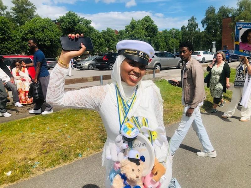 En leende ung kvinna i vit klänning och studentmössa står utomhus och håller upp en telefon, med gosedjur och medaljer hängande från halsen. Människor och grönska syns i bakgrunden.