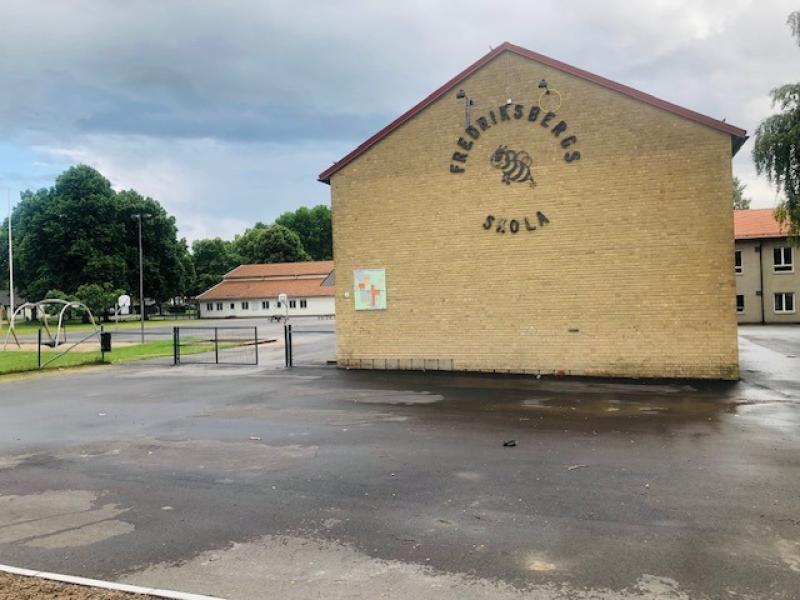 En gul skolbyggnad av tegel med skylten ”Fredriksbergets Skola” står bredvid en tom, asfalterad lekplats en molnig dag.