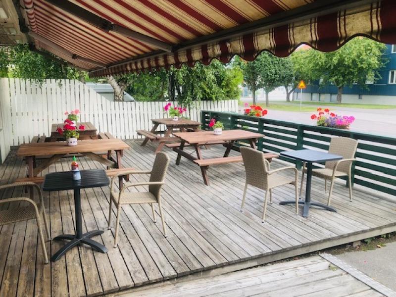 En uteplats med trägolv, picknickbord och stolar under ett randigt markistak, dekorerad med blommor i krukor. I bakgrunden syns ett vitt staket och grönska.