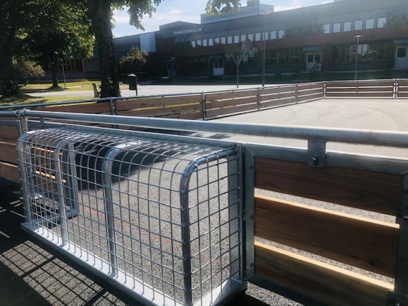 Ett inhägnat område av trä och metall med en skyddande trådbur, beläget utanför en modern byggnad med skylten ”TRANSTORP SKOLAN”. I bakgrunden syns träd och trottoar.