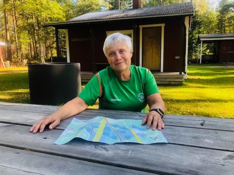 En äldre kvinna i en grön tröja sitter vid ett träbord utomhus och tittar på en karta. I bakgrunden syns en liten röd stuga och träd.