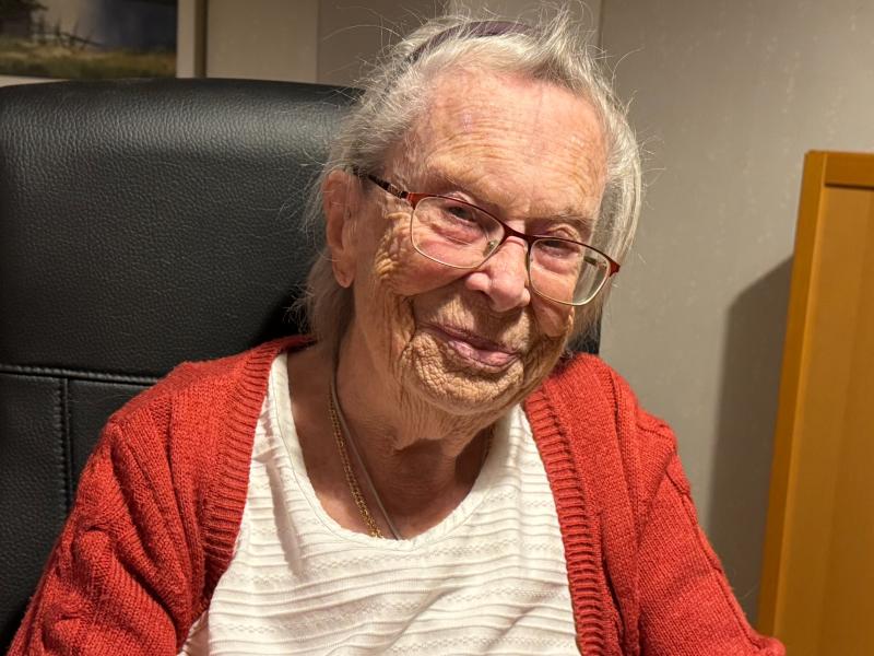 En äldre kvinna med glasögon och röd kofta sitter i en svart stol och ler mot kameran.