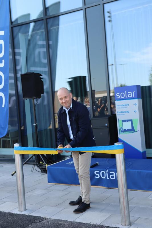 En man står utomhus och klipper av ett blått och gult band vid ett evenemang framför en byggnad med skyltar och banderoller med texten ”solar” i bakgrunden.